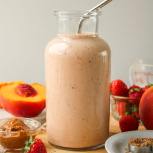 strawberry peach smoothie in a glass jug with a metal straw and ingredients on the cutting board around it