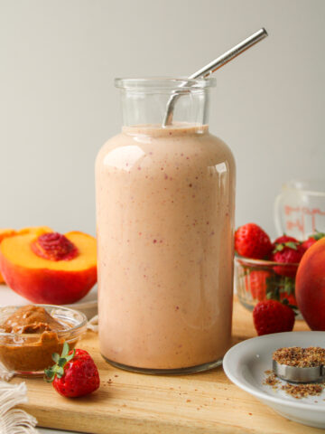 strawberry peach smoothie in a glass jug with a metal straw and ingredients on the cutting board around it