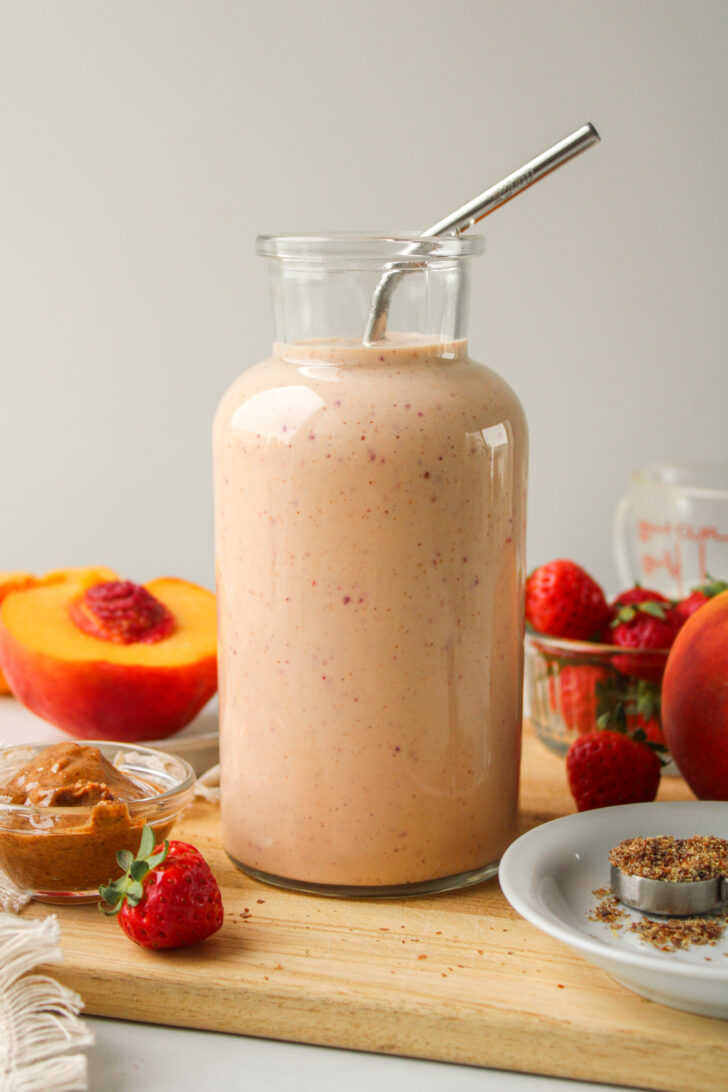 strawberry peach smoothie in a glass jug with a metal straw and ingredients on the cutting board around it