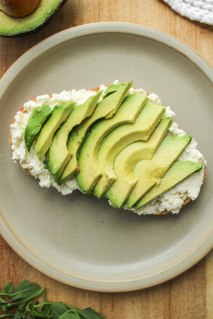 ricotta toast with slices of avocado on top on a ceramic plate on top of a wooden cutting board
