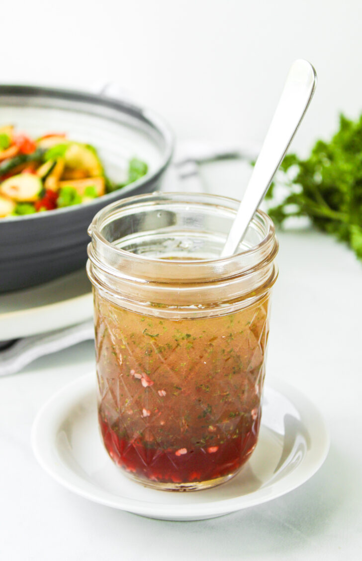 angled shot of red wine vinegar salad dressing in a glass mason jar with a metal spoon in it on top of a small white plate