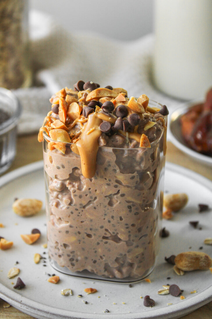 angled shot of snickers overnight oats in a glass container on a grey ceramic plate, topped with peanut butter, peanuts and mini chocolate chips