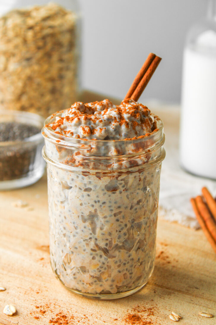 cinnamon roll overnight oats with a cinnamon stick in it in a glass mason jar on a wooden cutting board