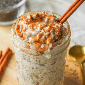 close up angled shot of cinnamon roll overnight oats with a cinnamon stick in it in a glass mason jar on a wooden cutting board