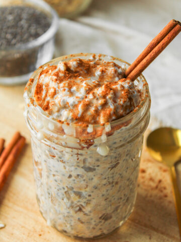 close up angled shot of cinnamon roll overnight oats with a cinnamon stick in it in a glass mason jar on a wooden cutting board