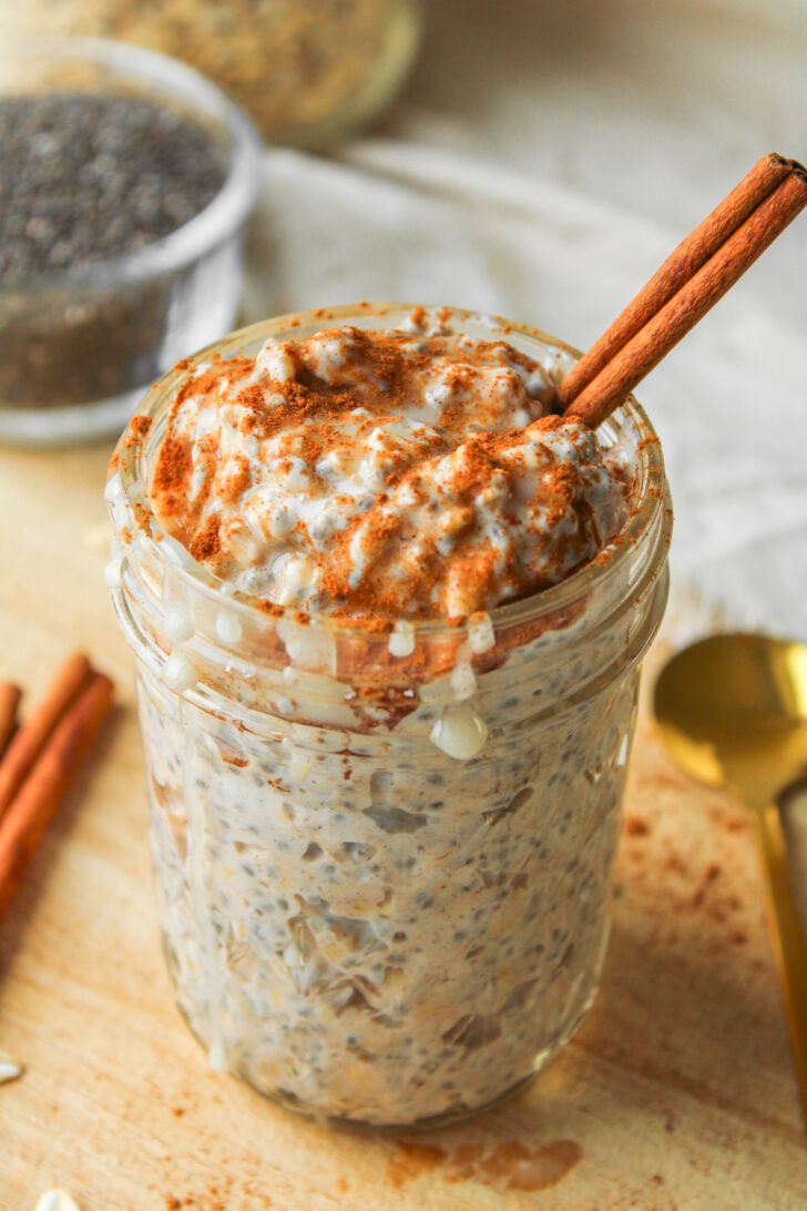 close up angled shot of cinnamon roll overnight oats with a cinnamon stick in it in a glass mason jar on a wooden cutting board