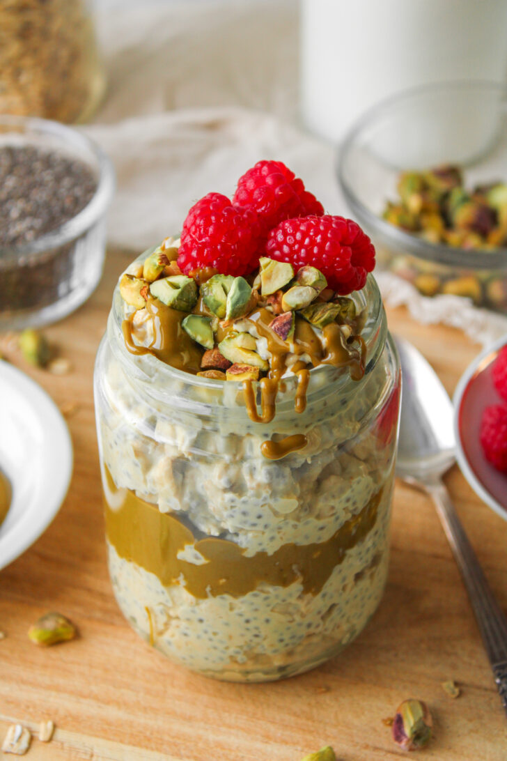 close up shot of pistachio overnight oats in a glass jar topped with pistachio butter, chopped pistachios and fresh raspberries