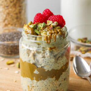 pistachio overnight oats in a glass jar topped with pistachio butter, chopped pistachios and fresh raspberries
