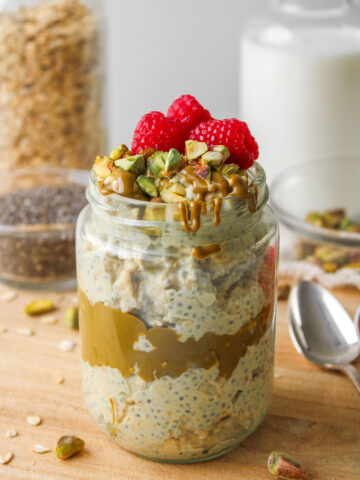 pistachio overnight oats in a glass jar topped with pistachio butter, chopped pistachios and fresh raspberries