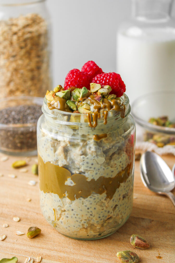 pistachio overnight oats in a glass jar topped with pistachio butter, chopped pistachios and fresh raspberries