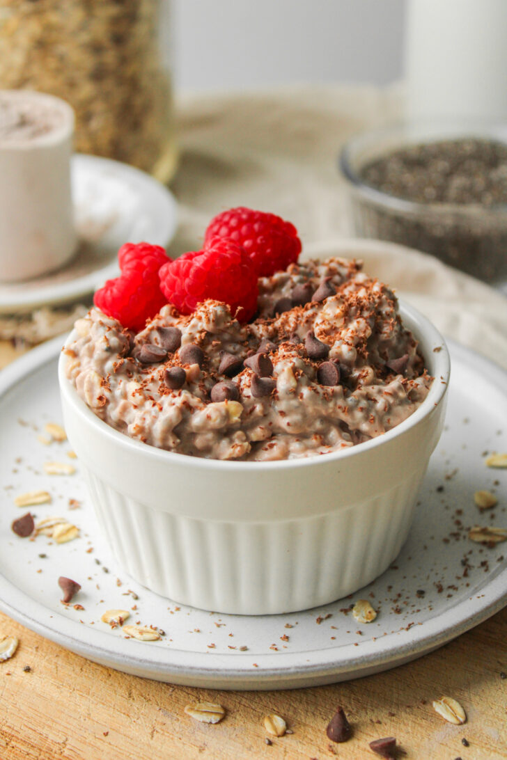 chocolate protein overnight oats, topped with chocolate chips and raspberries in a white ramekin dish on top of a white ceramic plate
