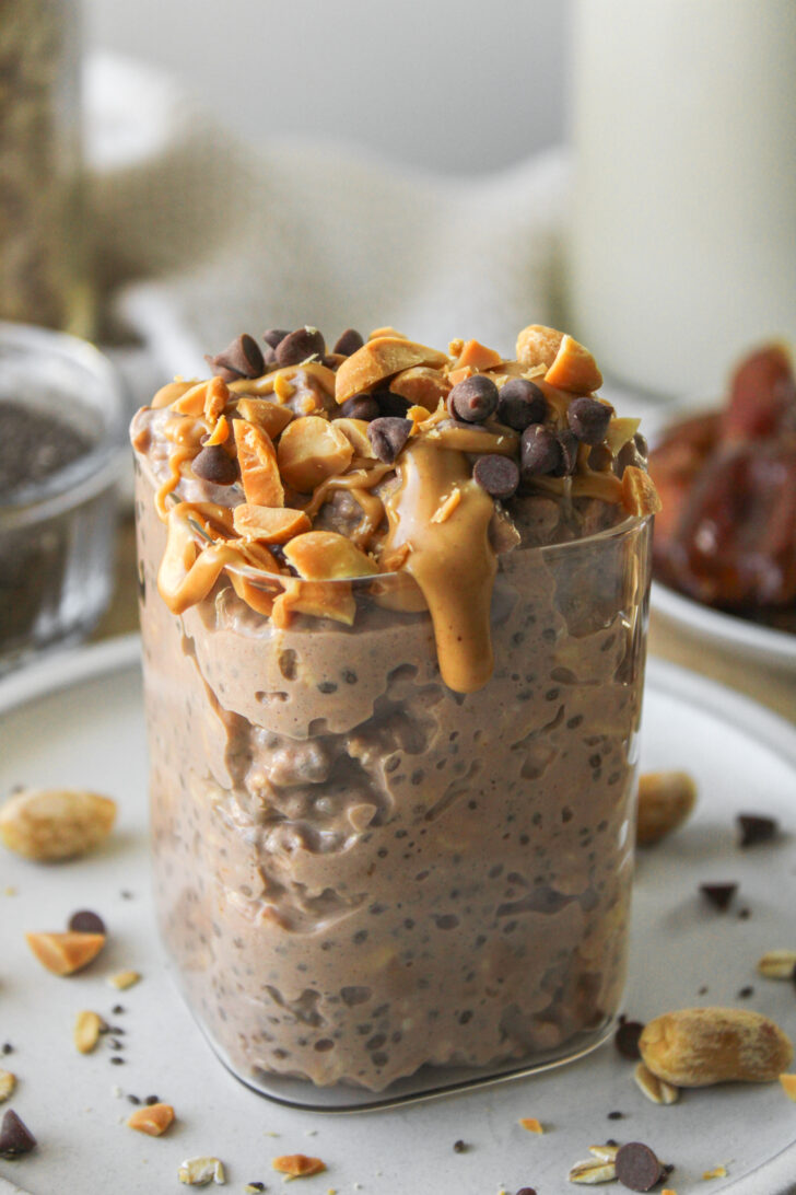 close up shot of snickers overnight oats in a glass container on a grey ceramic plate, topped with peanut butter, peanuts and mini chocolate chips