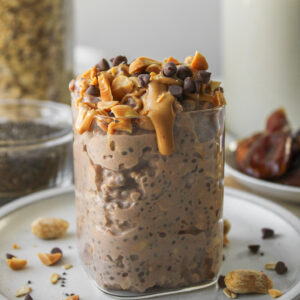 snickers overnight oats in a glass container on a grey ceramic plate, topped with peanut butter, peanuts and mini chocolate chips