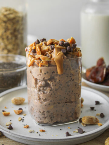 snickers overnight oats in a glass container on a grey ceramic plate, topped with peanut butter, peanuts and mini chocolate chips