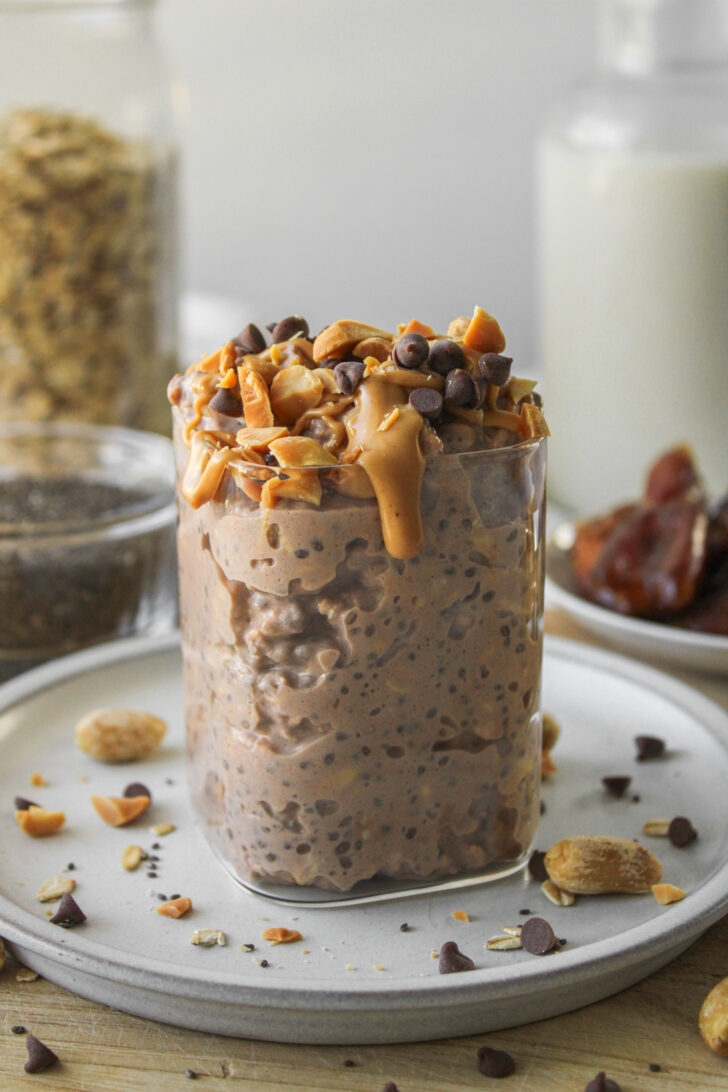 snickers overnight oats in a glass container on a grey ceramic plate, topped with peanut butter, peanuts and mini chocolate chips