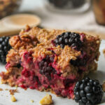 close-up of a square of blackberry baked oatmeal on a grey ceramic plate with ingredients in the background