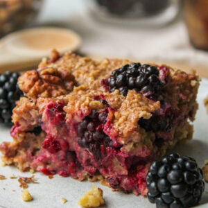 close-up of a square of blackberry baked oatmeal on a grey ceramic plate with ingredients in the background