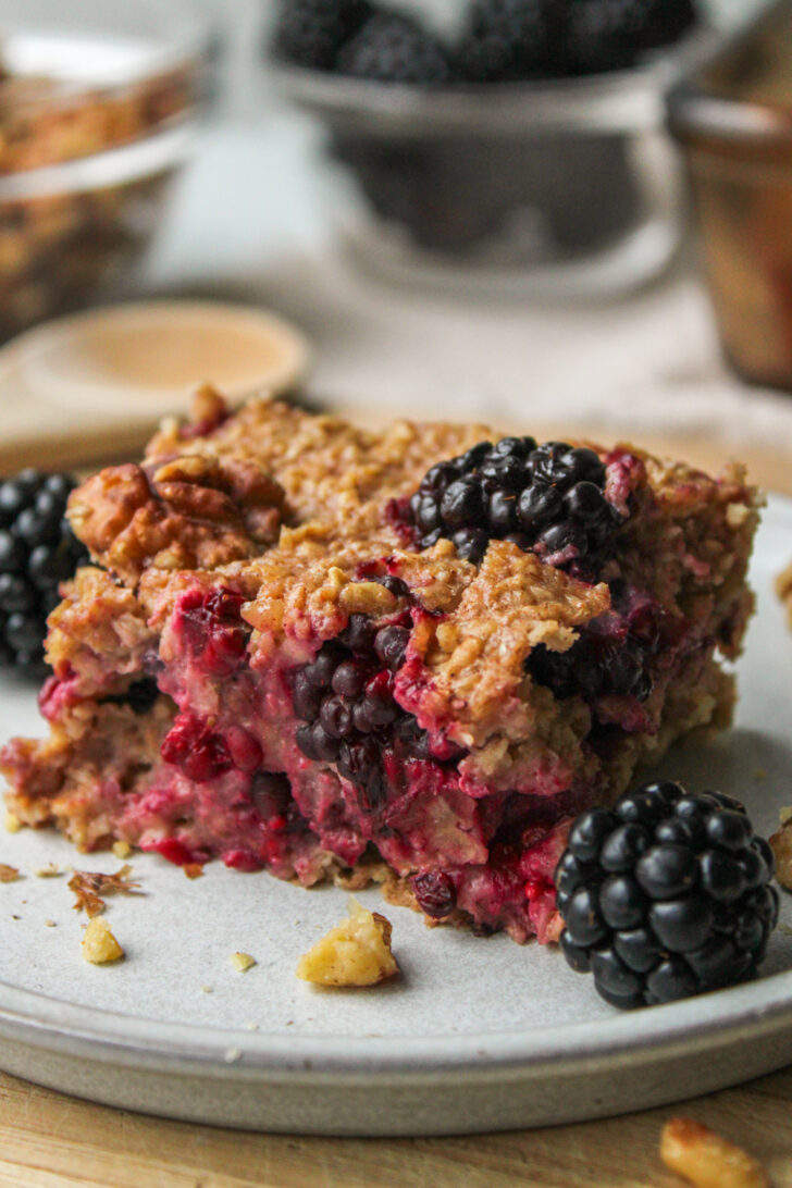 close-up of a square of blackberry baked oatmeal on a grey ceramic plate with ingredients in the background