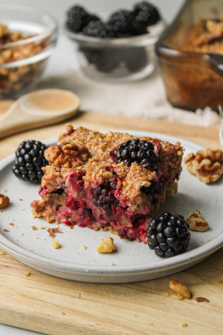 a square of blackberry baked oatmeal on a grey ceramic plate with ingredients in the background