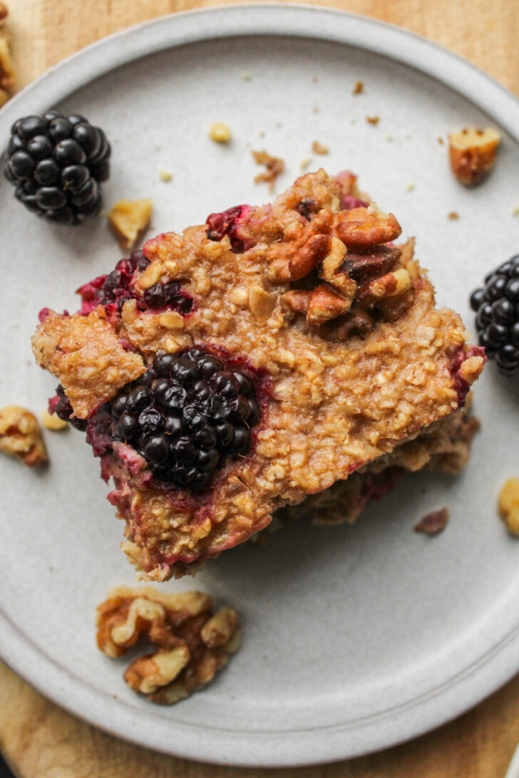 overhead shot of a square of blackberry baked oats on a grey ceramic plate with ingredients in the background