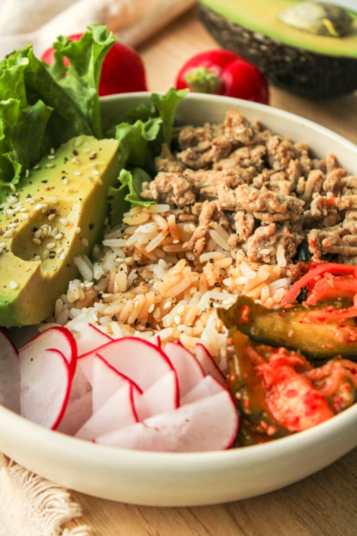 high protein kimchi rice bowls in a white ceramic bowl on top of a wooden cutting board and ingredients behind